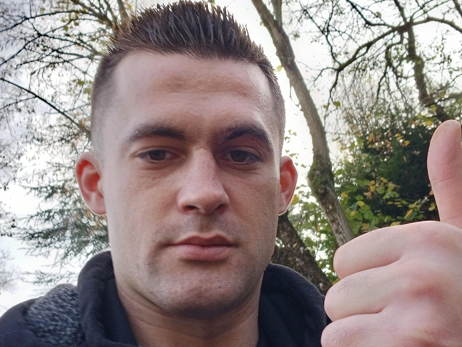 Matt69s, short brown spiked hair, fair skin, giving a thumbs-up, serious expression, wearing a black hoodie, outdoor setting, visible trees in background, slightly blurry, middle-aged dude.