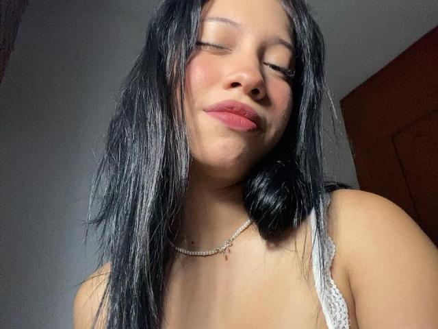 ImANaughtyBebe with black, wet-looking hair, eyes closed, pink lips, small tits, wearing a white lace top, and a delicate necklace, smiling slightly.