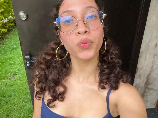ImANaughtyBebe with curly brown hair, wearing blue tank top, gold hoop earrings, and reflective blue glasses, pouting her full lips. Her brown skin is smooth and she's outdoors.