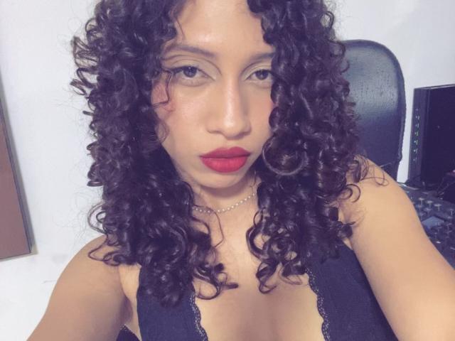 ImANaughtyBebe has curly black hair, red lipstick, and is wearing a black lace top that reveals her small tits. She's looking at the camera with a serious expression.