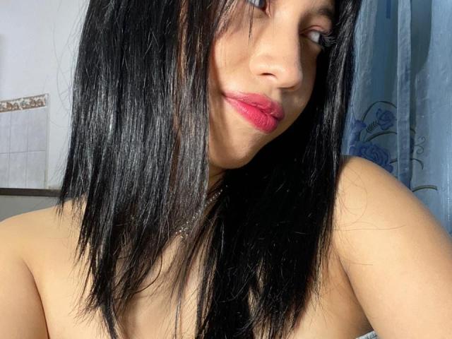 ImANaughtyBebe has straight black hair, pink lipstick, and a slight smile, looking to the side. She's topless with bare shoulders, showing smooth, light brown skin.