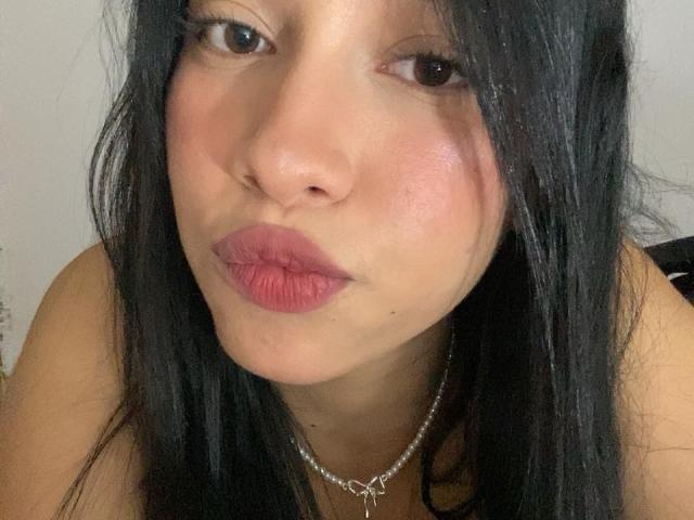 ImANaughtyBebe with puffy pink lips, dark straight hair, and a shiny necklace, giving a pouty, slightly smug facial expression. Her skin is lightly flushed.