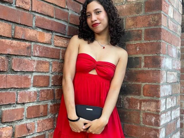 ImANaughtyBebe with curly black hair, red strapless dress, small boobs, holding black clutch, standing against red brick wall, wearing red lipstick and necklace.