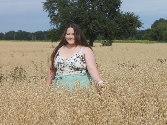 DaniellaGraceZZ stands in a golden wheat field, her long brown hair flowing. She's wearing a white floral tank top and a light blue skirt. A large tree is in the background. Her fair skin contrasts with the yellowish wheat. The sky is clear with a hint of blue. She's smiling, arms slightly raised. The field stretches out, with green trees in the distance. Her relaxed, confident posture dominates the image.