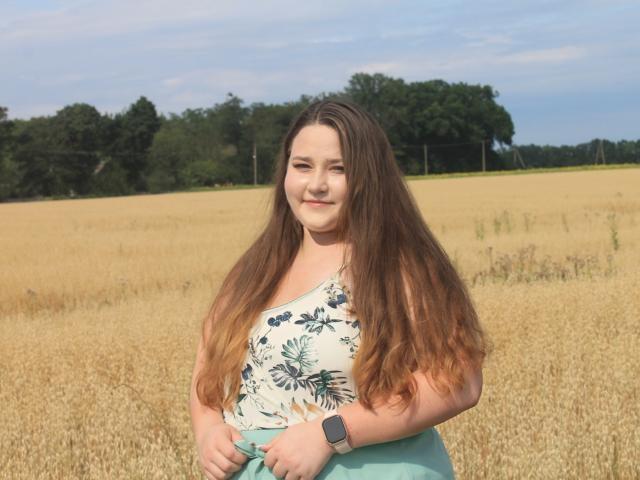 DaniellaGraceZZ stands in a golden wheat field, her long, wavy brown hair cascading over her shoulders. She wears a white floral tank top and a light blue skirt, with a black smartwatch on her left wrist. She has a subtle smile, light makeup, and a relaxed, natural look. The sky is blue with scattered clouds, and a line of green trees is in the distant background. The composition is balanced, with DaniellaGraceZZ centered and the field extending into the horizon. The texture of her hair contrasts with the smoothness of her top.