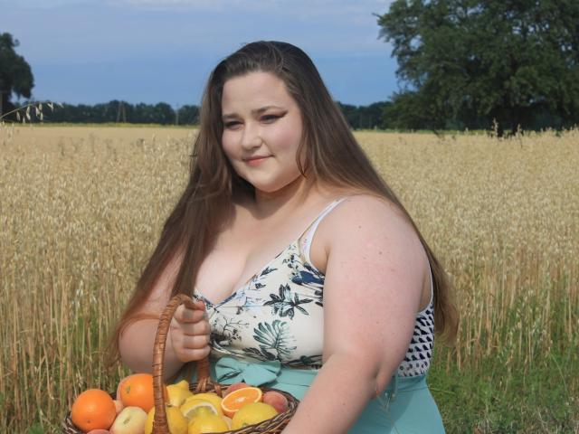 DaniellaGraceZZ, a plus-sized woman with long brown hair, sits in a wheat field. She wears a white floral tank top with a blue pattern and a mint green skirt. She holds a wicker basket filled with oranges, apples, and a peach. Her light skin contrasts with the golden wheat. The background shows a clear blue sky and green trees. Her subtle smile and relaxed posture convey a sense of calm. The image has a natural, candid feel.