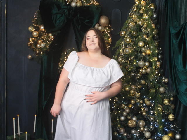 DaniellaGraceZZ stands in front of a decorated Christmas tree with golden ornaments and lights. She wears a white, short-sleeved dress, slightly holding her belly. Her brown hair is loose, and she has a neutral expression. Dark green velvet curtains frame the scene. Several white candles are on a green surface at the bottom. The composition uses rich, contrasting colors and textures. The image focuses on DaniellaGraceZZ against the festive, illuminated background.