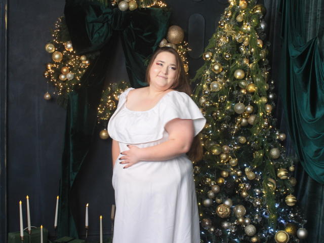 DaniellaGraceZZ, a plus-sized woman with long brown hair, stands in front of a decorated Christmas tree. She wears a white, short-sleeved dress, and her right hand rests on her slightly protruding belly. The Christmas tree, adorned with gold and silver ornaments and twinkling lights, is to her right. Several lit candles are at the tree's base. Dark green velvet curtains frame the scene. The image has a soft, warm light.
