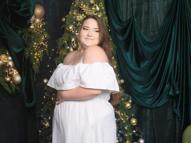 DaniellaGraceZZ with big ass, full tits, and a cute smile, wearing an off-shoulder white dress, standing in front of a Christmas tree with green drapes.