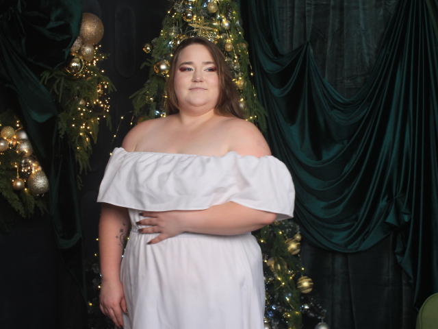 DaniellaGraceZZ, plus-size with pale skin, brown hair, off-shoulder white dress, hand on belly, standing in front of decorated Christmas tree, green velvet drapes, soft smile, subtle tattoo.