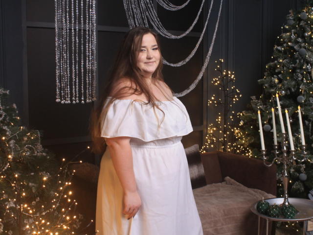 DaniellaGraceZZ stands in a dimly lit room. She has long brown hair, fair skin, and a soft smile. She wears an off-shoulder white dress with a yellow overlay. Her right arm is slightly raised. Behind her, a decorated Christmas tree with lights, a brown sofa, and a candleholder are visible. The room has black walls and string lights. She looks relaxed and confident. The composition is balanced with warm, soft lighting.
