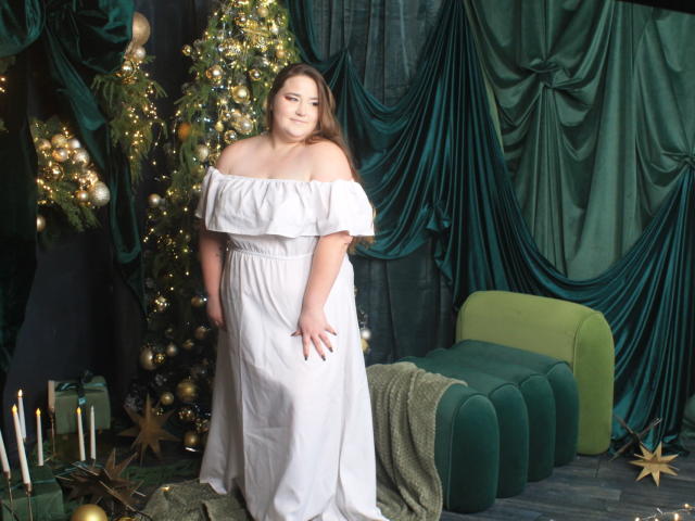DaniellaGraceZZ stands in a festive room wearing a white off-shoulder dress. She has light skin, brown hair, and a slight smile. A green velvet chaise lounge is to her right, and a decorated Christmas tree with gold and white ornaments is behind her. Dark green drapery hangs in the background. She has one hand on her dress, and there are candles and star ornaments on the floor. The photo has a watermark in the bottom right corner.
