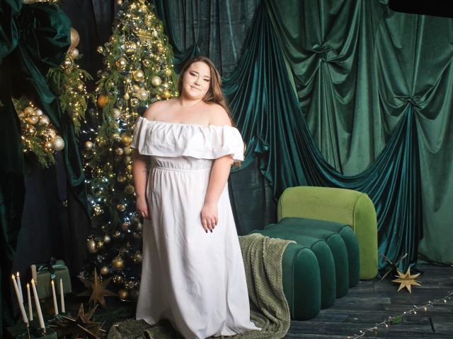 DaniellaGraceZZ stands in a green, velvety room wearing a white off-shoulder dress. She has long brown hair and a calm expression. A decorated Christmas tree with gold ornaments and lights is to her left, with a green velvet chaise and draped green fabric in the background. A green blanket is on the chaise, and candles and star ornaments are on the floor. The image has a festive, elegant vibe.
