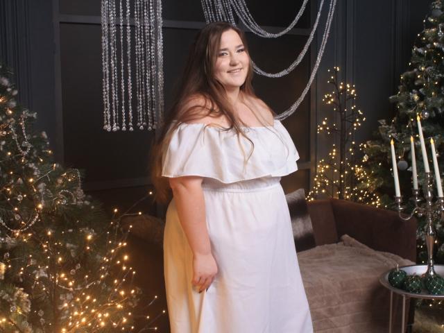 DaniellaGraceZZ with long brown hair, smiling, wearing an off-shoulder white dress, standing in front of Christmas trees with twinkling lights, and a candleholder on the right.