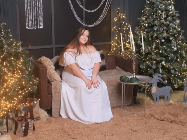 DaniellaGraceZZ, fat and cute, sitting on brown sofa in white off-shoulder dress, long brown hair, smiling, surrounded by Christmas trees, candles, and fairy lights.