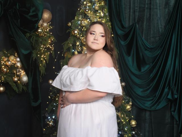 DaniellaGraceZZ with large tits, wearing a white off-shoulder dress, standing in front of a Christmas tree with green drapes, looking down with a slight smile.
