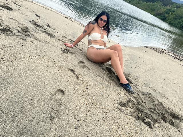 StellaBeckker with tits in a white bikini, sitting on sandy beach, legs crossed, tattoo on left arm, black hair, sunglasses, blue flip-flops, water in background.
