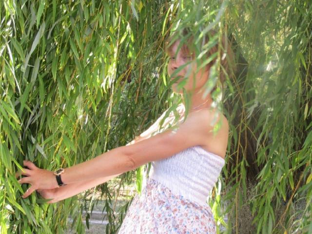 Lorraine Rux, con un top blanco sin tirantes y una falda estampada con flores, de pie en medio de bambú verde, con el brazo derecho extendido, una pulsera negra, y la luz del sol filtrándose entre las hojas..