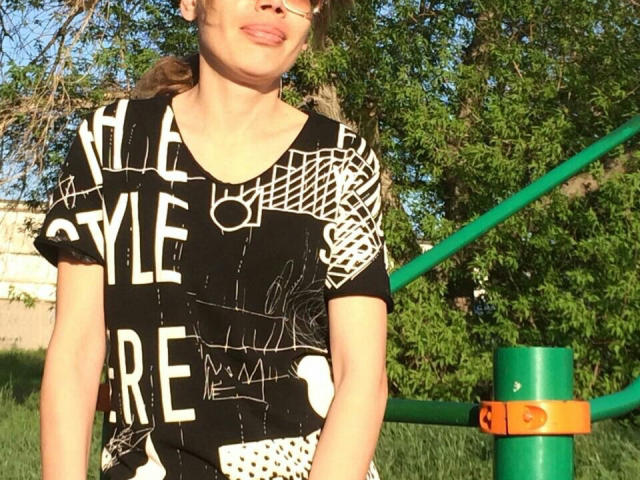 Lorraine Rux, com os lábios franzidos, vestindo um vestido com padrão preto e branco, tem os braços ao lado do corpo, em pé perto de uma estrutura de metal verde e laranja, em um dia ensolarado..