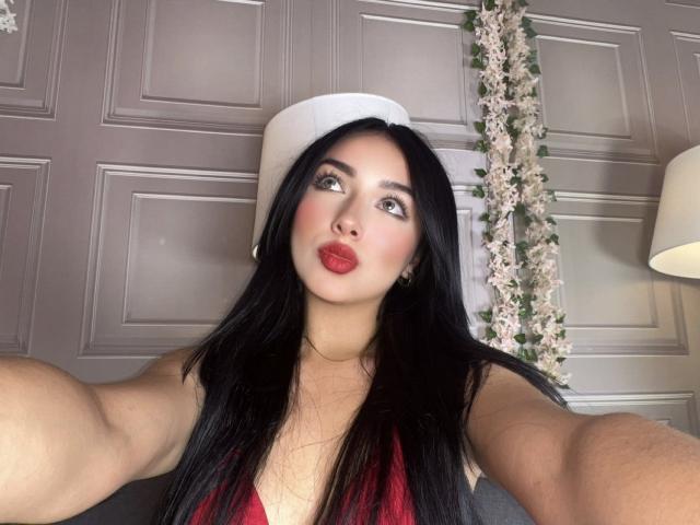 LillyTaylor with long black hair, red lipstick, and red top, pouting her plump ass at the camera, wearing a white hat, in a selfie.