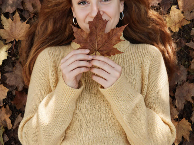 RedHurricane lies on a bed of brown and yellow fallen leaves. She holds a large, brown maple leaf close to her smiling face with both hands. She has long, wavy red hair and wears a thick, yellow knit sweater. Her earrings are small and silver. The texture of the sweater is ribbed, contrasting with the smooth, veined maple leaf. The image is centered on RedHurricane's upper body, with the leaf and her hands in the foreground. The leaves around her create a natural, textured background.