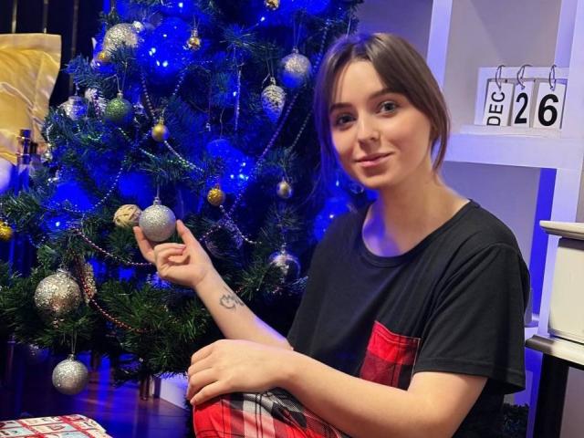 SofiaLuxury, a cute chick with brown hair, small tits, and a tattoo on her right arm, holds a glittery Christmas ornament, wearing a black and red plaid shirt.