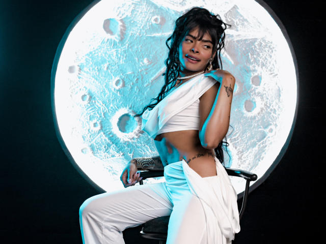 EmmaEbony with long black dreadlocks, wearing a white crop top and pants, sitting on a chair, pulling down her top to reveal her small tits, moon backdrop, smiling.