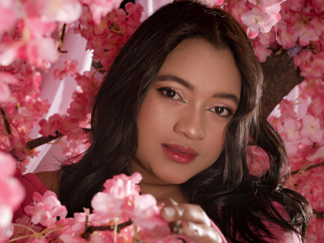 Il viso di SaritaCute, circondato da fiori di ciliegio rosa, con capelli scuri e ondulati, occhi marroni e rossetto rosa, guarda direttamente la fotocamera, mostrando un'espressione dolce e sensuale..