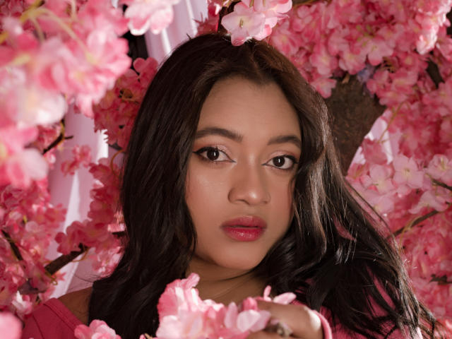 SaritaCute with dark wavy hair, pink lipstick, and brown eyes, surrounded by pink cherry blossoms, looking straight at the camera, her face centered in the shot.