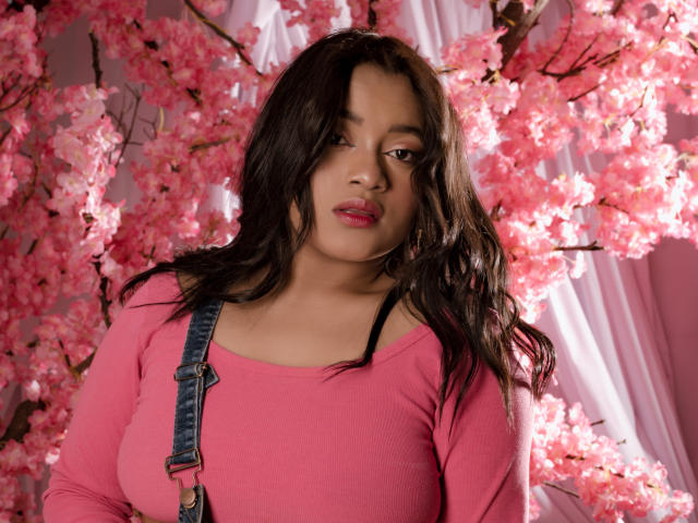 Sarita, niedlich mit einem rosa Kirschblütenhintergrund, trägt ein rosa Oberteil, Jeansriemen, dunkles, welliges Haar, rosa Lippenstift, leicht geschminkte Lippen, mittelgroße Brüste, weiches Licht, süß, attraktiv, selbstbewusst..