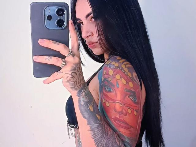 SafiraGreludinha with long black hair, tattoos on her right arm, taking a selfie with a grey phone, showing her colorful, detailed arm tattoo and black bra.