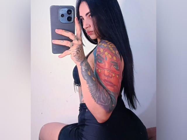 SafiraGreludinha with long black hair, tattoos on her arm, wearing a black dress, taking a mirror selfie, showing off her butt, holding an iPhone, looking sexy.