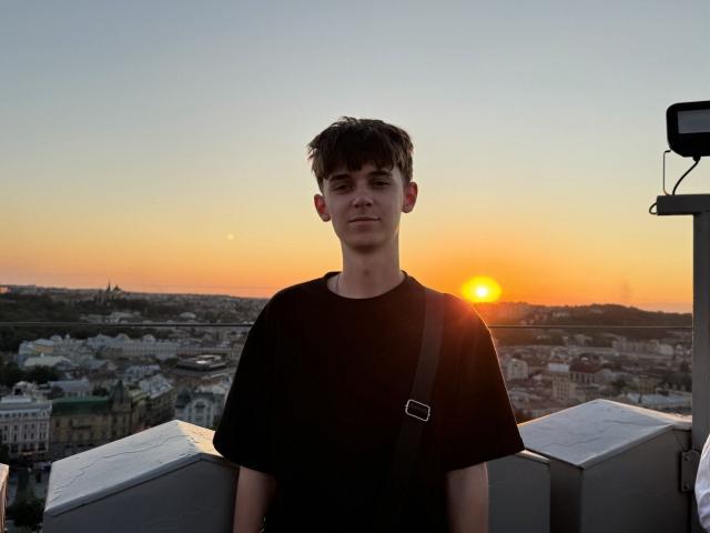 Un ragazzo, SweetDude, con capelli castani corti, indossa una maglietta nera e una cinghia a tracolla attraversata sul petto, in piedi su un tetto al tramonto, con il sole alle sue spalle e la città sullo sfondo..