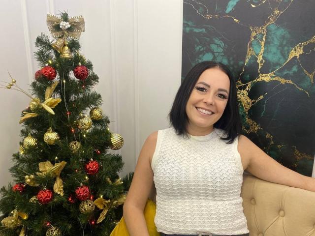 Samantha Reider, avec une coupe de cheveux courte noire, une robe blanche en dentelle, souriante, assise à côté d'un sapin de Noël décoré avec des ornements dorés et rouges, sur une chaise jaune..