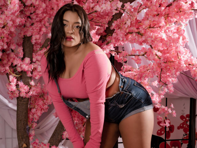 Sarita, niedlich mit einem pinken Crop-Top, das ihre Brüste zeigt, und Jeansshorts, lehnt sich nach vorne an rosa Kirschblüten, blickt mit rotem Lippenstift, braunen Haaren und mittlerem Hautton in die Kamera..
