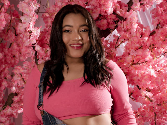 Sarita, com um visual adorável com flores de cerejeira rosa, vestindo uma blusa cropped rosa, macacão jeans, sorrindo, mostrando a parte de cima do abdômen e com seios de tamanho médio. Ela tem cabelo escuro ondulado e batom rosa..