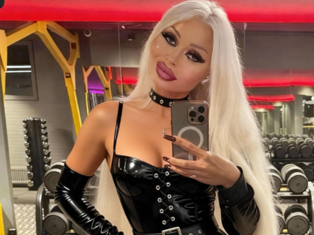 RoxyRoyale with long white hair, black leather corset, and gloves, holding a phone, wearing a black choker, in a gym, showing her tits and a confident smile.