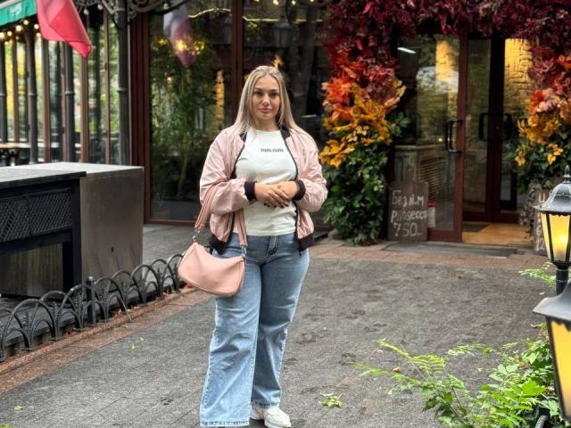 Dawn, met blond haar, een wit T-shirt, blauwe jeans, een roze jas en een roze handtas, staat op een geplaveide stoep, houdt iets in haar handen en kijkt vooruit..