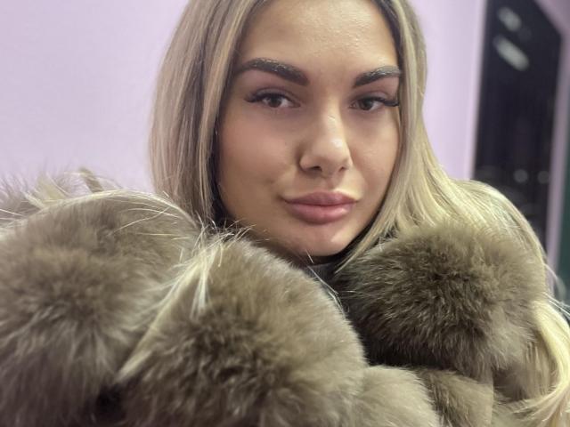 Dawn, met blond haar, dikke wenkbrauwen en roze lippen, draagt een bontjas en kijkt met een lichte glimlach in de camera. Haar borsten zijn gedeeltelijk bedekt door de jas..