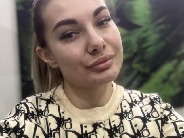 DawnBelinda with light skin, brown eyebrows, and blonde hair in a ponytail, wearing a black-and-white patterned sweater, has a small pimple on her right cheek, smiling softly.