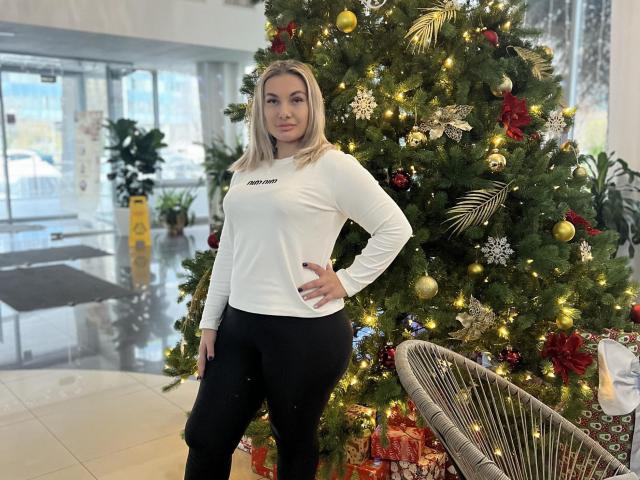 DawnBelinda stands in front of a decorated Christmas tree. She's wearing a white long-sleeve shirt with "AMAZON" text and black leggings. Her blonde hair is straight and falls to her shoulders. The tree is adorned with gold, red, and silver ornaments, and small lights. A wicker chair with a patterned cushion is to the right. DawnBelinda's right hand is on her hip, and she looks directly at the camera. The room is brightly lit with large windows in the background.