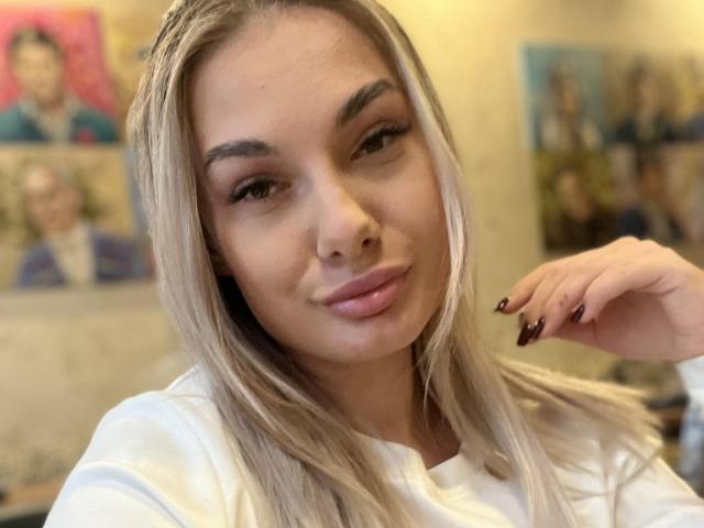 Dawn Belinda hat blondes Haar, dicke Augenbrauen und zwinkert leicht, mit einem kleinen Lächeln. Sie trägt ein weißes Hemd und hat dunkelrote Fingernägel. Sie sieht sowohl sexy als auch niedlich aus..