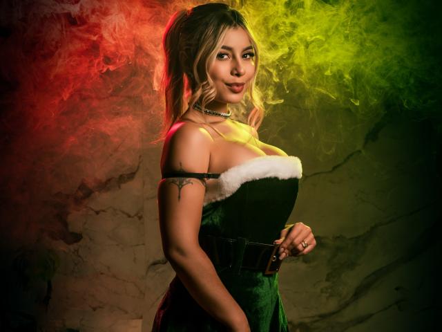 AmaraRicci, avec ses cheveux blonds attachés en queue de cheval, portant une robe en velours vert avec des finitions en fourrure blanche, montrant sa poitrine, debout devant de la fumée colorée, souriant avec assurance..