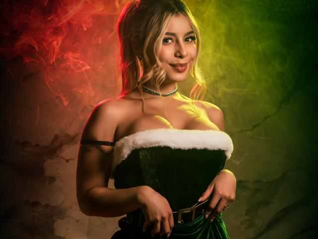 AmaraRicci, com cabelo loiro, vestido verde de veludo sem alças, detalhes de pele branca, cinto e colar, sorrindo, com fumaça vermelha e verde, mostrando seus seios grandes e uma tatuagem sexy..