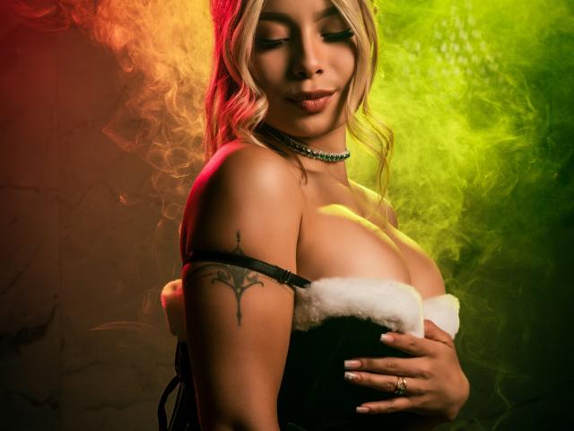AmaraRicci, met grote borsten, blond haar en een tatoeage op haar arm, draagt een zwart-witte top met imitatiebont, groene en rode rook, en een groene kralenketting..