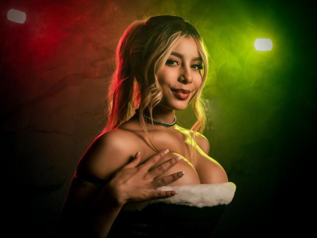 AmaraRicci, avec des couettes blondes, des lumières vertes et rouges, portant un haut noir sans bretelles avec de la fausse fourrure blanche, cachant ses grandes seins avec sa main, souriante, et portant un collier..