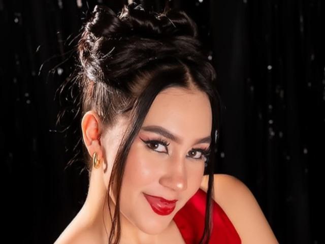 CelesteRusso with dark, wavy hair in a messy bun, red lipstick, gold earrings, and a red dress, smiling softly at the camera.