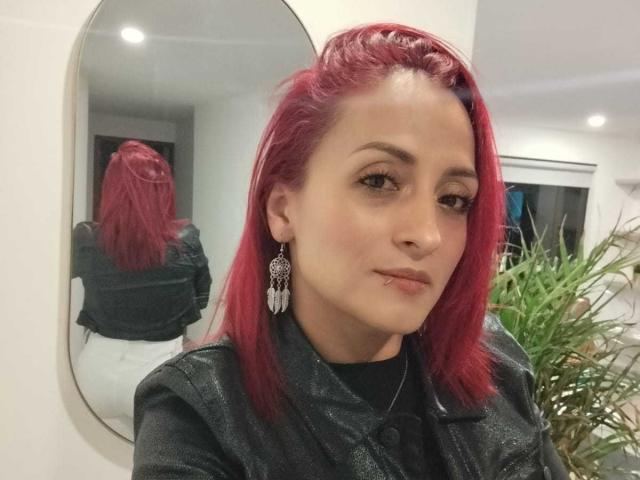 AmandaSolery with red hair, wearing a black jacket, has a nose ring and feather earrings, reflected in a mirror. Her ass is in the background mirror reflection.