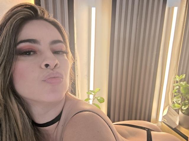 ChristineBray with pouty lips, brown hair, pink eyeshadow, black choker, and black bra, showing her tits and butt, standing in front of vertical light strips.