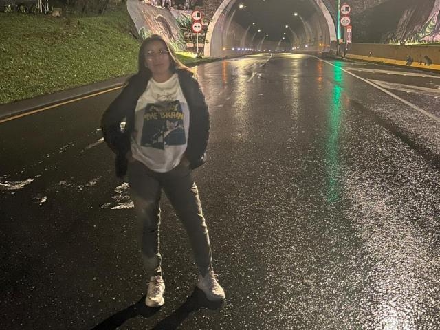 KathyLoveWild standing in a dark, wet tunnel at night, wearing a white "The Rolling Stones" shirt, black jacket, gray pants, and white sneakers, with a confident, relaxed stance.